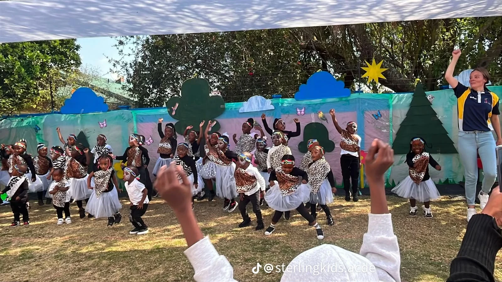 Academy children and a teacher take part in a lively outdoor performance beneath a canopy.