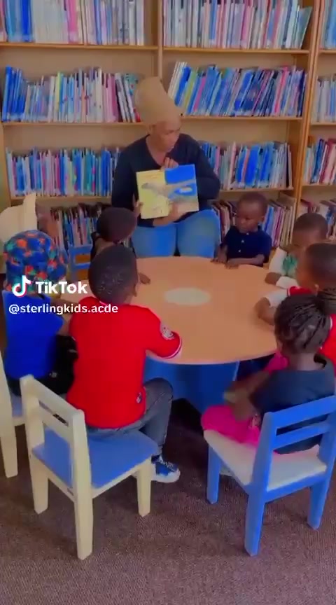 Children sit around a table listening to a story in front of shelves of books.